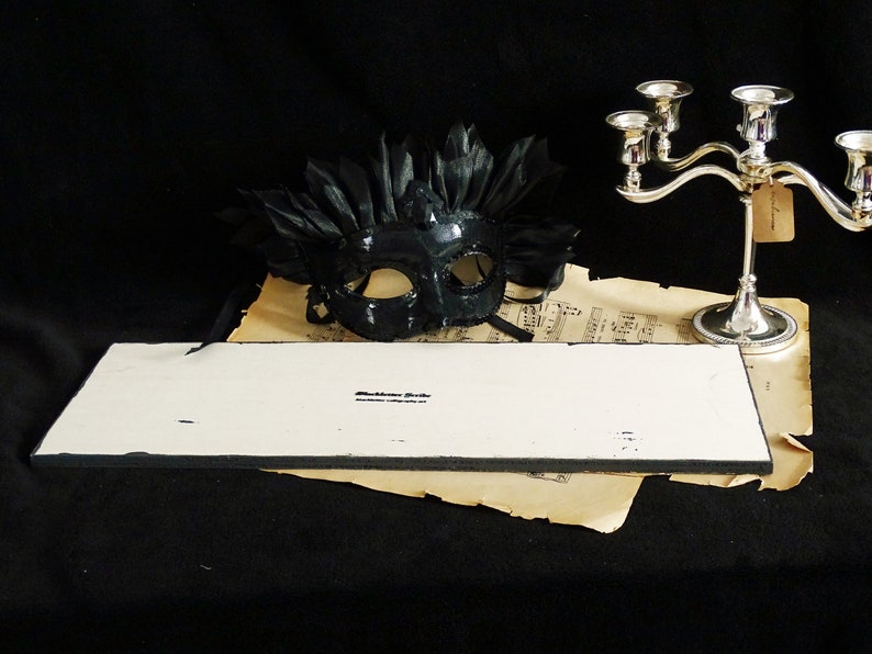 May include: A white rectangular sign with black edges. The sign has the text "Blackstone Gifts" printed on it. The sign is sitting on top of a piece of sheet music with a black masquerade mask with feathers on top of the sheet music.