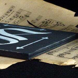 May include: A black wooden sign with white calligraphy and decorative accents. The sign is resting on a sheet of music.