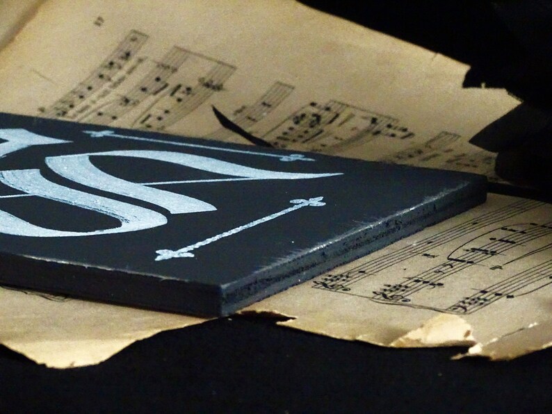 May include: A black wooden sign with white calligraphy and decorative accents. The sign is resting on a sheet of music.