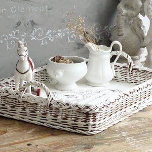 Può includere: Un vassoio di vimini bianco con manici, contenente una brocca, una ciotola e una statuina di cavallo in ceramica bianca. Il vassoio è appoggiato su una superficie di legno, con uno sfondo dipinto ed elementi decorativi.