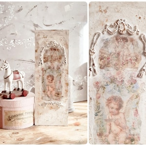 May include: A rectangular wooden wall plaque with a distressed white finish, featuring a central transfer print of a cherub and roses, framed by raised white decorative moulding. The plaque displays a soft, aged aesthetic with visible texture.