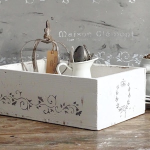 Könnte beinhalten: Eine rechteckige, weiß lackierte Holzkiste mit Used-Look, verziert mit grauen, schablonenartigen Blumenranken und einem Kronen-Emblem an der Seite. Die Kiste enthält ein metallenes, kronenförmiges Drahtgestell, ein kleines weißes Keramik-Kännchen.