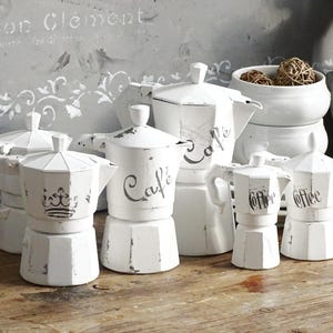 May include: A collection of white, distressed-finish coffee pots of various sizes. Some pots have the word "Cafe" or "Coffee" stenciled on them. The background includes a decorative wall panel with French text and a white horse figurine.