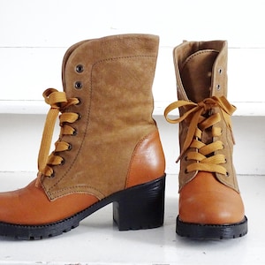 May include: A pair of mid-calf boots featuring a tan leather toe and heel counter, a brown suede upper shaft, and mustard yellow laces. The boots have a black block heel and a thick, lugged black rubber sole with metal eyelets for the lacing system.