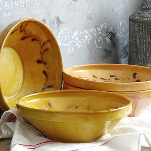 Könnte beinhalten: Eine Sammlung gelber Keramikschalen im rustikalen Stil. Die Schalen haben eine glänzende Oberfläche und dunkelbraune dekorative Akzente. Sie sind auf einem weißen Tuch angeordnet, was auf eine Küche oder einen Essbereich hindeutet.