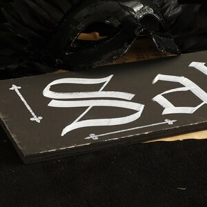 May include: A black wooden sign with the word "Sale" written in white calligraphy. The sign is decorated with small white arrows and fleur-de-lis.