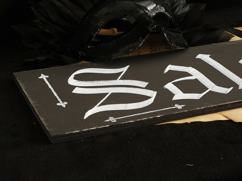 May include: A black wooden sign with the word "Sale" written in white calligraphy. The sign is decorated with small white arrows and fleur-de-lis.
