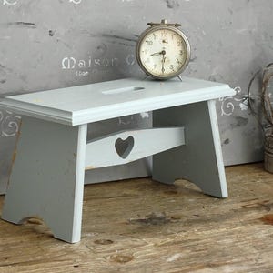 Puede incluir: Un taburete de madera de color gris claro con un recorte en forma de corazón en el soporte central y una ranura rectangular en la parte superior. El taburete presenta un acabado desgastado con vetas de madera y bordes gastados en las patas.