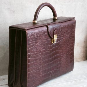 May include: A dark brown briefcase with a textured crocodile-embossed pattern. The bag features a single rounded top handle, a fold-over flap with a gold-toned metal push-lock closure, and a structured rectangular shape with expandable side gussets.