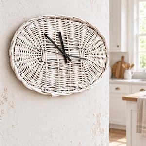Puede incluir: Un reloj de pared ovalado con un marco de mimbre blanco trenzado y una esfera con textura de tejido de cesta. El reloj tiene tres manecillas finas y rectas de metal negro situadas en el centro, que contrastan con el material claro del tejido.
