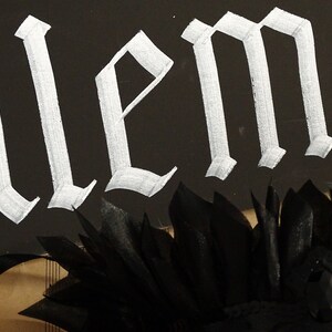 May include: A black background with the word "alem" written in white gothic script. A black feather boa is at the bottom of the image.