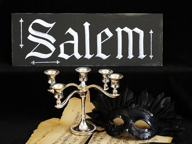 May include: A black chalkboard sign with the word "Salem" written in white calligraphy. Below the sign is a silver candelabra with five candle holders, a black masquerade mask with sequins, and a sheet of music.