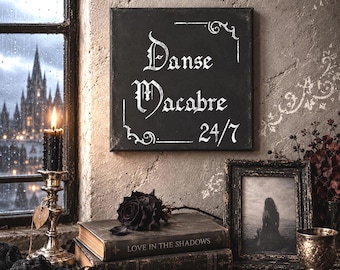 May include: A square black sign with white text reading Danse Macabre 24/7 in a gothic font, featuring decorative corner flourishes. The sign is displayed on a textured wall above a stack of books, a black rose, and a framed photograph.