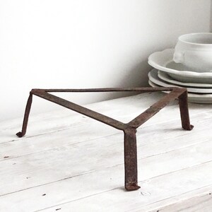 Puede incluir: Un salvamanteles triangular de metal con un acabado oscuro y oxidado y tres patas resistentes. El marco está construido con tiras de metal planas unidas en las esquinas, proporcionando una base estable para utensilios de cocina.