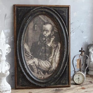 Op de afbeelding: Een zwart ovaal ingelijst portret van een man met een baard, die een donker jasje en een witte kraag draagt. De lijst heeft een gouden rand.