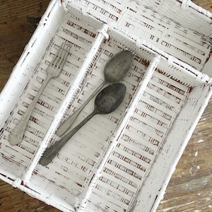May include: A white painted wicker cutlery tray with three compartments. A silver fork and two spoons rest inside the tray. The tray sits on a weathered wooden surface, showcasing a rustic aesthetic.