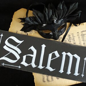 May include: A black chalkboard sign with the word "Salem" written in white gothic script. The sign is resting on a sheet of music with a black masquerade mask on top.