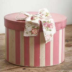 Puede incluir: Una caja de almacenamiento cilíndrica y redonda con rayas verticales de color rosa y crema. La tapa es de color rosa sólido y está rematada con un lazo de tela decorativo que muestra un patrón floral de rosas rosas y hojas verdes sobre un fondo crema.