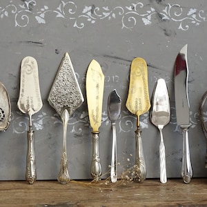 Op de afbeelding: Een verzameling van acht metalen serveerbestekken, waaronder taartscheppen, smeermessen en een mes. De set bevat diverse zilver- en goudkleurige afwerkingen, gedetailleerde reliëfpatronen en sierlijke handvatten, opgesteld in een rij.