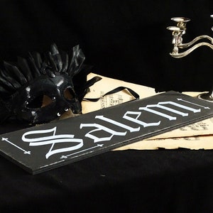 May include: A black wooden sign with the word "Salem" written in white script. The sign is sitting on top of a piece of sheet music. A black masquerade mask with feathers is in the background.