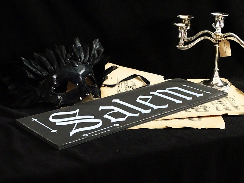 May include: A black wooden sign with the word "Salem" written in white script. The sign is sitting on top of a piece of sheet music. A black masquerade mask with feathers is in the background.