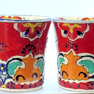 May include: Two red ceramic drinking glasses with colorful floral and geometric patterns. The glasses have a vibrant design with orange, blue, yellow, and green accents. The rims are trimmed with a blue and yellow border. The glasses are likely handmade.