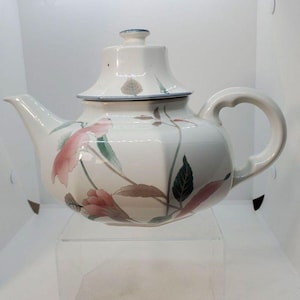 May include: A white ceramic teapot with a faceted body and a matching lid. The teapot features a painted botanical design with soft pink flowers, muted green leaves, and brown stems. It has a curved handle, a tapered spout, and a blue rim detail.