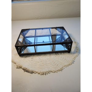 May include: A rectangular glass display box with a mirrored base and dark metal framing. The box features six internal compartments separated by thin glass dividers and decorative filigree metal corner accents on the hinged lid.