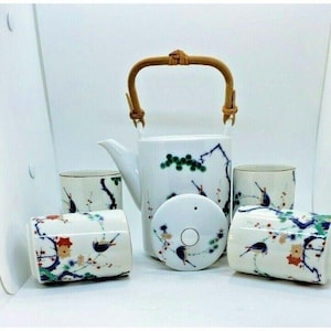 Puede incluir: Un juego de té de cerámica blanca con una tetera, tapa y cuatro tazas. La tetera tiene un asa de bambú. Cada pieza está decorada con un diseño de pájaros y flores en azul, verde y rojo. El juego está diseñado para servir té.