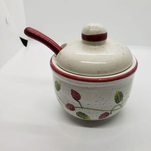 May include: A ceramic sugar bowl with a lid and spoon. The bowl is off-white with a red rim and a floral design. The lid has a red stripe. The spoon is red.