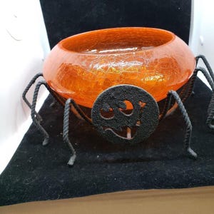 May include: A round orange crackle glass bowl supported by a black metal stand featuring eight thin, textured legs and a circular front emblem with a cutout design. The bowl has a translucent, webbed texture throughout its vibrant orange surface.
