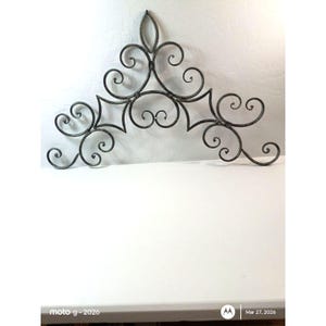 May include: A decorative metal wall art piece with a symmetrical design. The piece features swirling scrollwork and geometric shapes in a dark gray color. The design is mounted on a white surface.