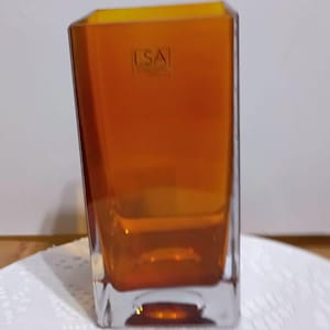 May include: An orange rectangular glass vase with a clear base. The vase has a slightly angled design and a square opening. The text "LSA" is visible on the front of the vase.