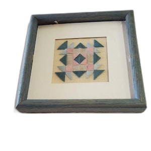 May include: A framed needlepoint artwork featuring a geometric design in shades of pink, teal, and gray. The artwork is set against a cream-colored background and encased in a gray wooden frame. The design is a square pattern.