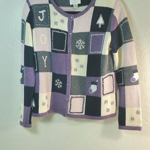 May include: A long-sleeve button-down cardigan featuring a patchwork design in shades of purple, black, and cream. The knit garment displays embroidered holiday motifs including snowflakes, stockings, a tree, and the letters J, O, and Y.