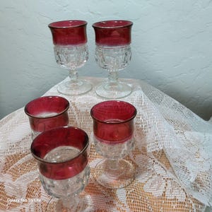 May include: Set of five clear glass stemmed drinking glasses with a ruby red rim. The glasses are on a white lace tablecloth.