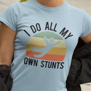 May include: A light blue t-shirt with a retro-style graphic that says "I do all my own stunts". The graphic features a stick figure doing a backflip in front of a sunset.
