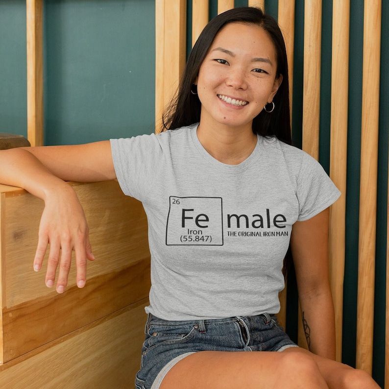 May include: A grey t-shirt with a black graphic of a periodic table element. The element is labelled "Fe" for Iron and the atomic number is 26. The text "Female" is written next to the element box with the words "The Original Iron Man" underneath.