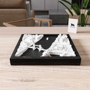 May include: A white 3D model of London, England, featuring the Tower Bridge and surrounding buildings, presented in a black wooden frame. The model is displayed on a wooden table. A white card with the text "CITY3D" is visible in the background.
