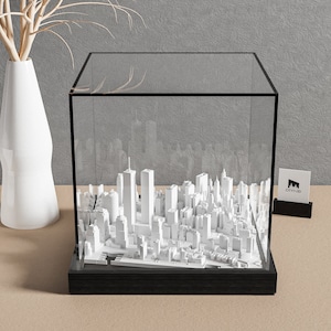 May include: A white 3D model of the New York City skyline, including the World Trade Center towers, is displayed in a clear glass cube with a black base. The cube is sitting on a light brown surface. The word "CITYMOD" is printed on a small black card in the background.