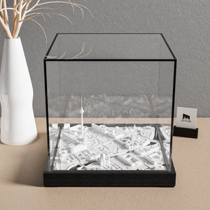 Pode incluir: Um modelo 3D branco de um horizonte urbano dentro de uma vitrine de vidro transparente com uma base preta. O modelo inclui edifícios, ruas e uma torre alta. A vitrine está sobre uma superfície marrom clara.
