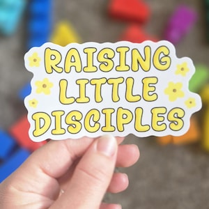 May include: A white sticker with the text "RAISING LITTLE DISCIPLES" in yellow, outlined in black. Small yellow flower graphics are scattered around the edges. The sticker is held by a hand.