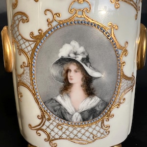 Op de afbeelding: Een witte porseleinen vaas met gouden accenten en een geschilderd portret van een vrouw met een witte hoed. De vaas heeft een ronde basis met drie gouden voetjes.