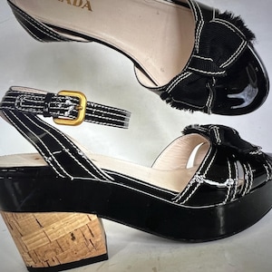 May include: Black patent leather platform sandals with a cork wedge heel and a white stitched bow detail. The sandals have an ankle strap with a gold buckle.