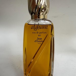 Op de afbeelding: Een doorzichtige glazen fles parfum met een gouden dop. De fles is gevuld met een gele vloeistof. Het etiket op de fles luidt "afghane eau de parfum 80° Jean d'Estrees PARIS".
