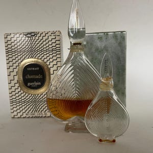 May include: Two glass perfume bottles with a diamond-shaped design. The larger bottle is filled with a brown liquid and has a gold label that reads "extrait charmade guerlain paris". The smaller bottle is empty and has a clear label that reads "charmade".