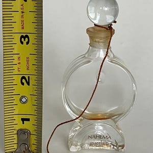 May include: A clear glass bottle of perfume with a round stopper and a brown string tied around the neck. The bottle is labeled "NAHEMA guerlain" and is sitting on a white surface.
