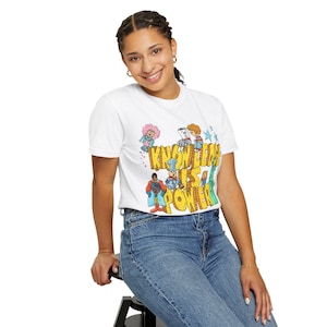 May include: White t-shirt with the text "KNOWLEDGE IS POWER" in yellow and colorful cartoon illustrations. The shirt features a crew neck and short sleeves. The design includes various cartoon characters and stars.