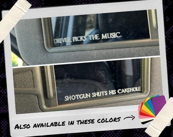 May include: Two white vinyl decals applied to car sun visor mirrors. The top decal reads "DRIVER PICKS THE MUSIC" and the bottom decal reads "SHOTGUN SHUTS HIS CAKEHOLE" in a bold, capitalised font. A colour swatch fan is shown in the bottom corner.