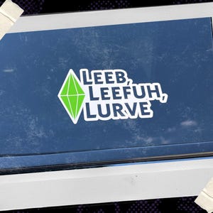 May include: A white sticker with a green diamond-shaped plumbob icon and the text LEEB, LEEFUH, LURVE in bold white letters with a dark blue outline, displayed on a dark blue surface within a white frame with tape accents at the corners.